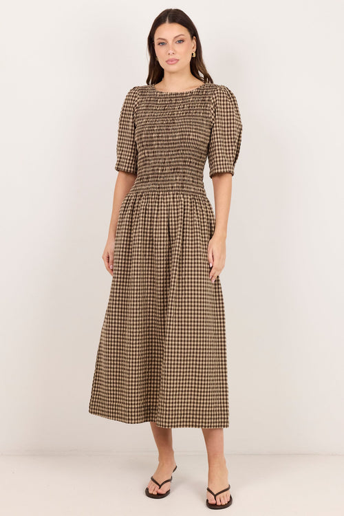 A woman modeling a brown and beige checkered midi dress with puff sleeves and a smocked bodice, standing against a plain off-white studio background.