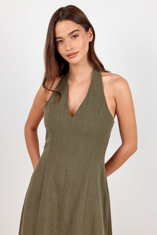 A woman modeling a sleeveless olive green halter-neck dress against a plain light gray studio background.