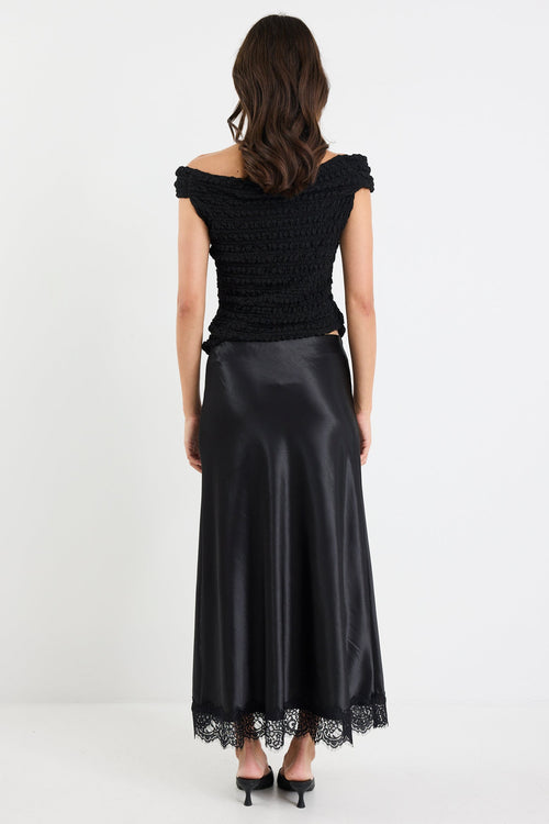 Woman wearing a black off-shoulder top and black satin skirt with lace hem on a white background