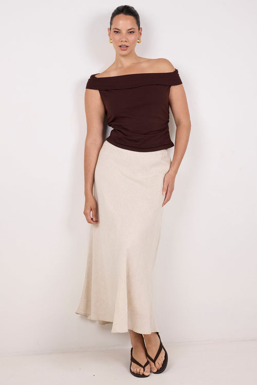 A woman modeling an off-the-shoulder dark brown top and a long cream skirt, paired with black strappy sandals, against a plain white studio background.