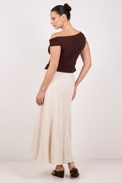 A woman modeling a dark brown off-the-shoulder top and a long cream-colored skirt, shown from a side-back angle against a plain white studio background.