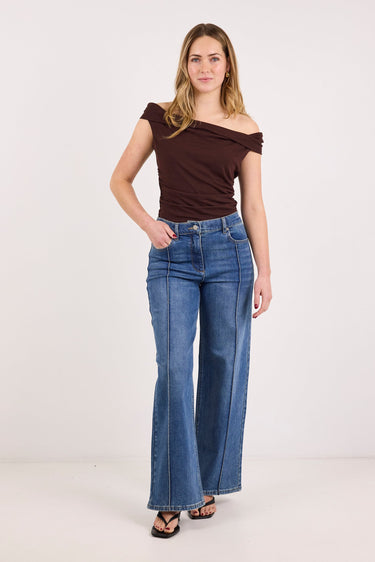 A woman modeling a dark brown off-the-shoulder top and blue wide-leg jeans against a plain white studio background.