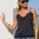 A woman wearing a black lace-trimmed camisole top and light-wash blue jeans, standing outdoors against a white wall and blue sky.
