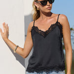 A woman wearing a black lace-trimmed camisole top and light-wash jeans, standing outdoors against a white wall under bright sunlight.