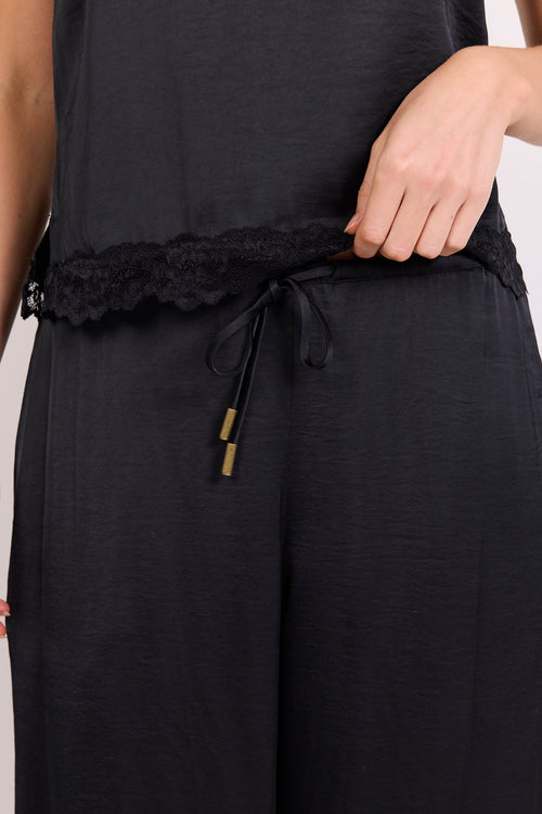 Close-up of a person wearing black wide-leg pants with a lace-trimmed drawstring waistband and a black sleeveless top, against a light neutral background.