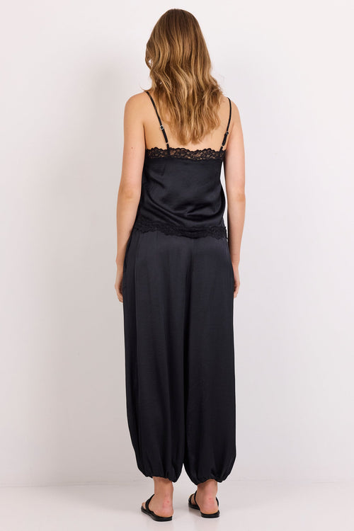 A woman viewed from behind wearing a black satin camisole with lace trim and matching black wide-leg harem pants, standing against a plain white studio background.