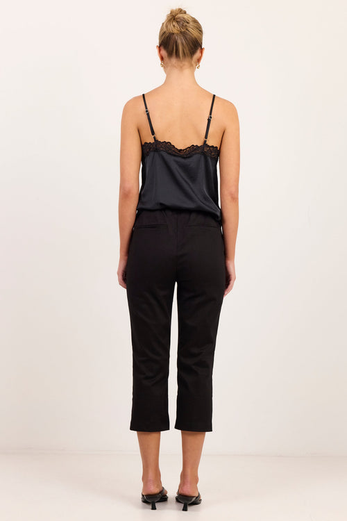 A woman viewed from behind wearing a black satin camisole with lace trim and black cropped trousers, standing against a plain white studio background.