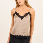 A woman modeling a beige camisole top with black lace trim along the neckline and hem, paired with black pants, against a plain light beige studio background.