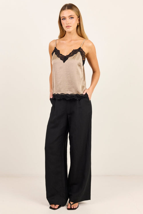 A woman modeling a beige camisole top with black lace trim and black wide-leg pants against a plain off-white studio background.