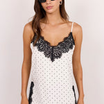 A woman modeling a white camisole top with black lace trim and black polka dots, paired with blue jeans, against a plain light background.