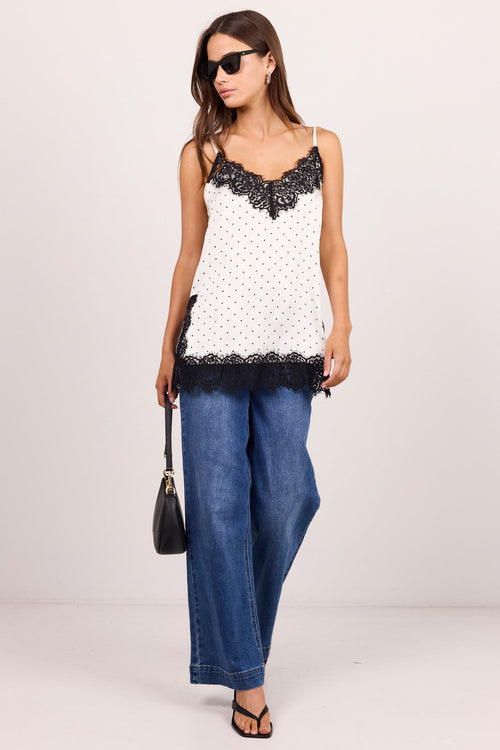 A woman modeling a white sleeveless top with black lace trim and black polka dots, paired with wide-leg blue jeans, black sandals, and a small black handbag, against a plain light gray studio background.
