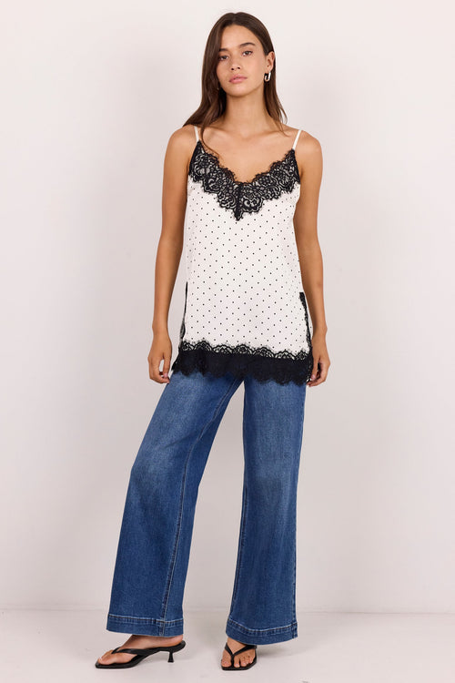 A woman modeling a white camisole top with black lace trim and blue wide-leg jeans against a plain white studio background.