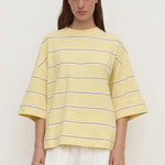 A model wearing a pale yellow oversized short-sleeve t-shirt with thin dark horizontal stripes, paired with white pants, against a plain off-white background.