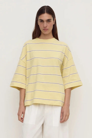 A model wearing a pale yellow oversized short-sleeve t-shirt with thin dark horizontal stripes, paired with white pants, against a plain off-white background.