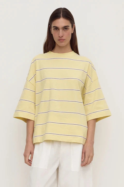 A model wearing a pale yellow oversized short-sleeve t-shirt with thin dark horizontal stripes, paired with white pants, against a plain off-white background.