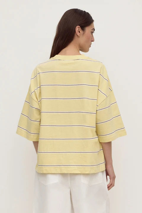 A woman shown from the back wearing a pale yellow short-sleeve striped T-shirt with thin dark horizontal stripes and white pants, photographed against a plain light background.