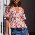 A woman wearing a short-sleeve wrap-style blouse with a red and pink floral print on a white background, paired with blue jeans, standing by a white door.