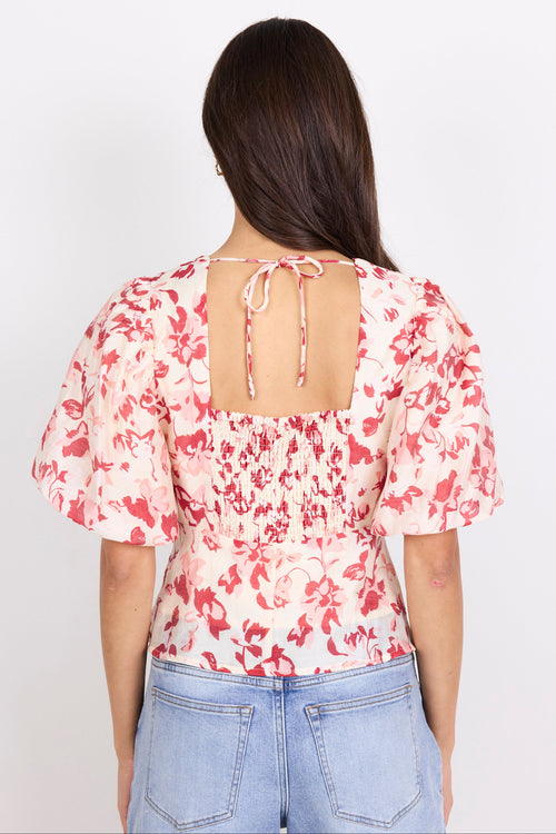 A woman viewed from the back wearing a short puff-sleeve top with a red floral print on a light pink background and a tie-back neckline, paired with light blue jeans against a plain white studio background.