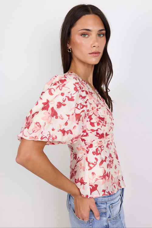 A woman modeling a short-sleeve blouse with a red and pink abstract floral print on a light cream background, paired with light blue jeans, against a plain white studio background.