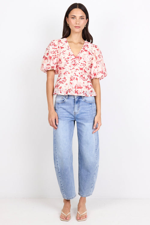 A woman stands against a plain white studio background wearing a short-sleeve floral blouse with puff sleeves, light blue jeans, and clear high-heel sandals.