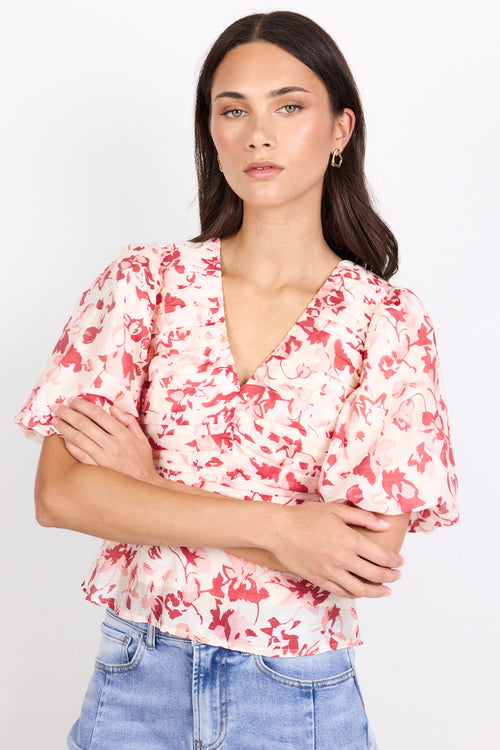 A woman wearing a short-sleeve wrap-style blouse with puff sleeves and a red floral print on a light pink background, paired with light blue jeans, against a plain white studio background.