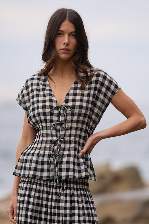 A woman modeling a matching black and white gingham check set consisting of a short-sleeve tie-front top and a long skirt, set against a blurred coastal background.