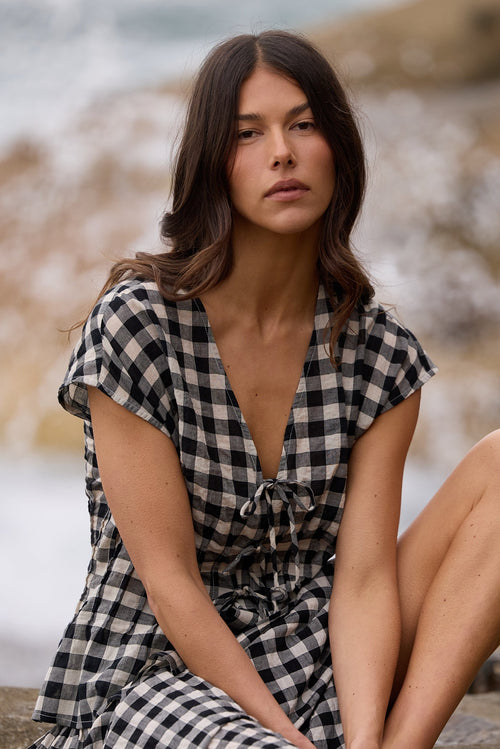 A woman sits on a large rock by the ocean, wearing a black and white gingham wrap dress with short sleeves and a tie at the neckline.