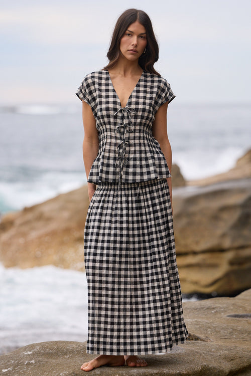 A woman stands barefoot on large rocks by the ocean, wearing a black and white gingham check peplum maxi dress with short cap sleeves and a V-neckline.