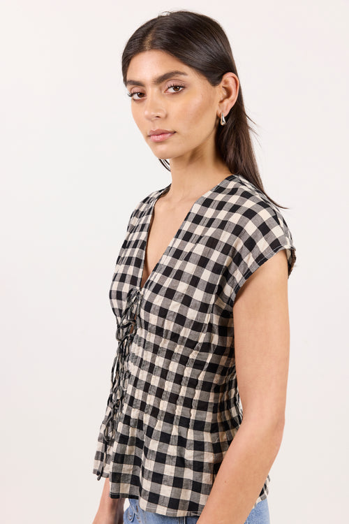 A woman wearing a black and beige gingham check wrap top with side tie details, paired with light blue jeans, photographed against a plain white studio background.