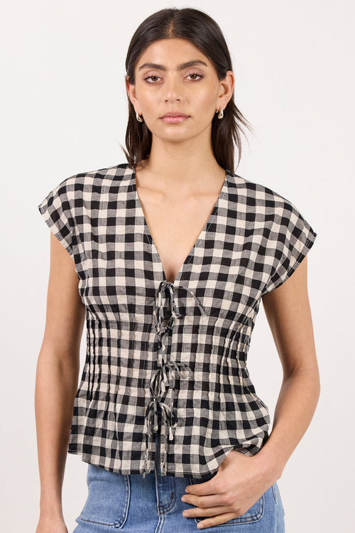 A woman modeling a black and white gingham checkered top with a deep V-neckline and front tie details, paired with light blue jeans, against a plain off-white studio background.