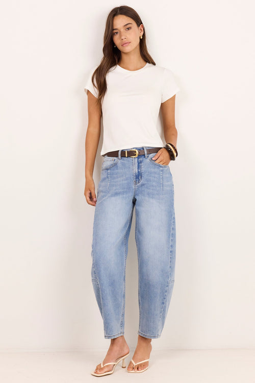 A woman modeling a pair of light-wash, wide-leg cropped jeans with a dark belt, paired with a white t-shirt and beige sandals, standing against a plain off-white background.