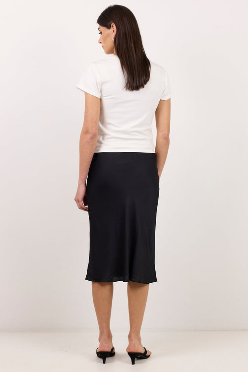 A woman is shown from behind wearing a white short-sleeve t-shirt and a black midi skirt, paired with black slingback heels, against a plain off-white studio background.
