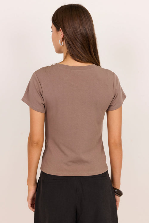 A woman viewed from the back wearing a short-sleeve crewneck t-shirt in a muted taupe-brown color, paired with black pants, against a plain off-white background.