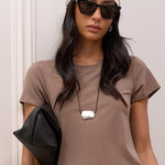 A woman stands indoors against a white paneled wall and wooden stairs, wearing a taupe short-sleeve cropped T-shirt, a black midi skirt, black sandals, sunglasses, a white patterned headscarf, and a black shoulder bag.