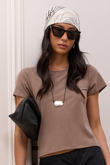 A woman stands indoors against a white paneled wall and wooden stairs, wearing a taupe short-sleeve cropped T-shirt, a black midi skirt, black sandals, sunglasses, a white patterned headscarf, and a black shoulder bag.