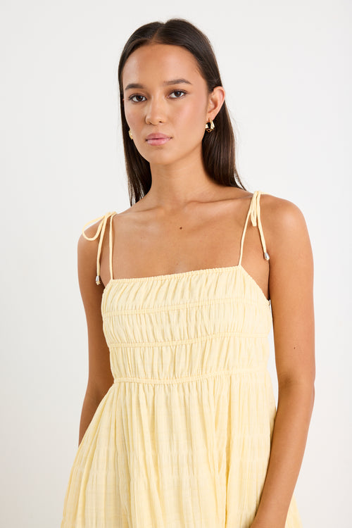Woman wearing a pale yellow shirred maxi dress with thin tie-up straps and a flowing tiered skirt, paired with neutral sandals.