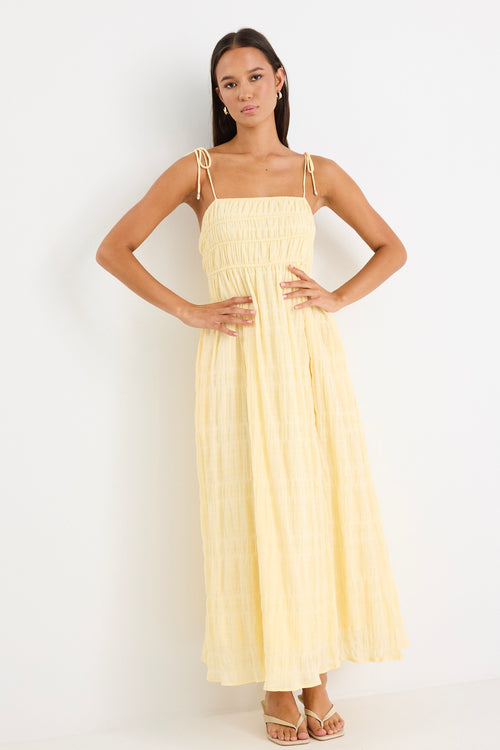 Woman wearing a pale yellow shirred maxi dress with thin tie-up straps and a flowing tiered skirt, paired with neutral sandals.