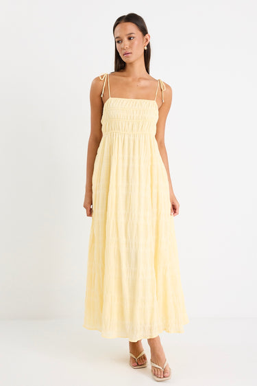 Woman wearing a pale yellow shirred maxi dress with thin tie-up straps and a flowing tiered skirt, paired with neutral sandals.