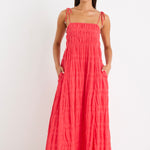 Woman wearing a poppy red shirred maxi dress with tiered detailing and tie-up straps, styled with white sandals.
