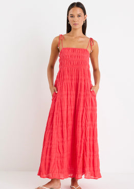 Woman wearing a poppy red shirred maxi dress with tiered detailing and tie-up straps, styled with white sandals.