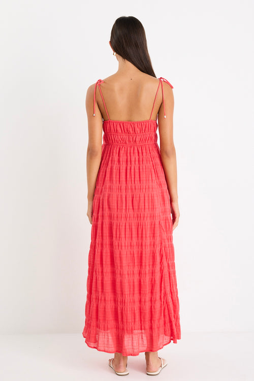 Woman wearing a poppy red shirred maxi dress with tiered detailing and tie-up straps, styled with white sandals.