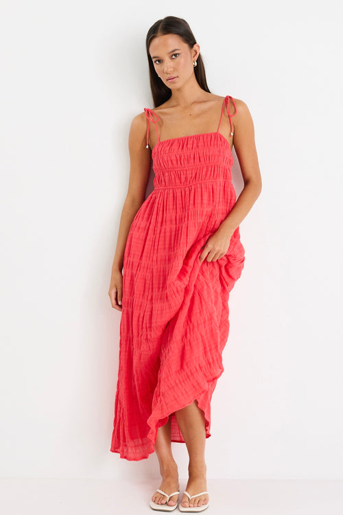Woman wearing a poppy red shirred maxi dress with tiered detailing and tie-up straps, styled with white sandals.