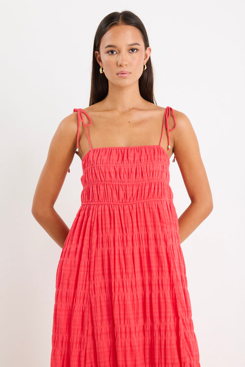Woman wearing a poppy red shirred maxi dress with tiered detailing and tie-up straps, styled with white sandals.