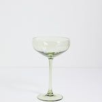 A single light green champagne coupe glass with a wide, shallow bowl, a slender stem, and a flat circular base, photographed against a seamless white studio background.