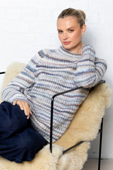 Woman sitting on a chair with a textured blanket wrapped around her, wearing a striped sweater and dark pants.