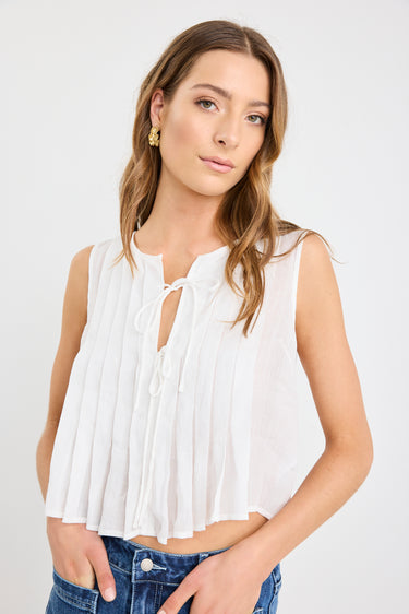 Woman wearing an ivory pleated sleeveless top with tie neckline, styled with high-waisted blue jeans.