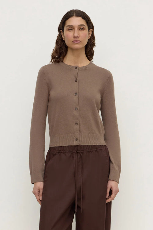 A woman modeling a taupe brown button-up cardigan sweater paired with dark brown drawstring trousers, shot against a plain light gray studio background.