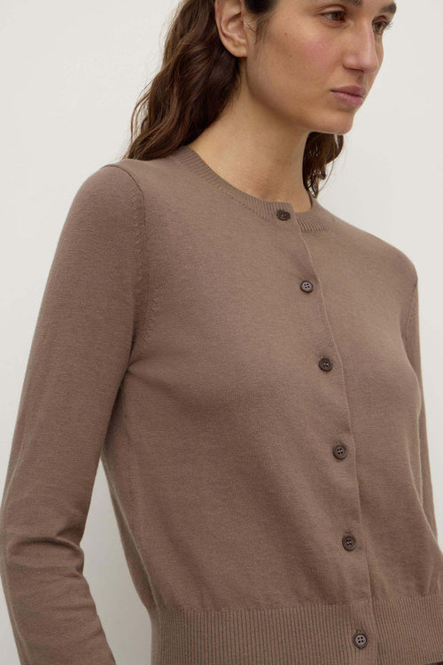 A close-up of a woman wearing a taupe brown long-sleeve button-up cardigan sweater against a plain light background.