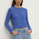A woman wearing a royal blue long-sleeve cropped button-front cardigan and light-wash blue jeans, standing against a plain light gray wall.