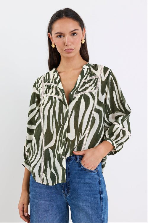 Woman wears a khaki and cream zebra-print shirt with lace inserts and pintuck detail, styled with gold earrings and blue jeans.